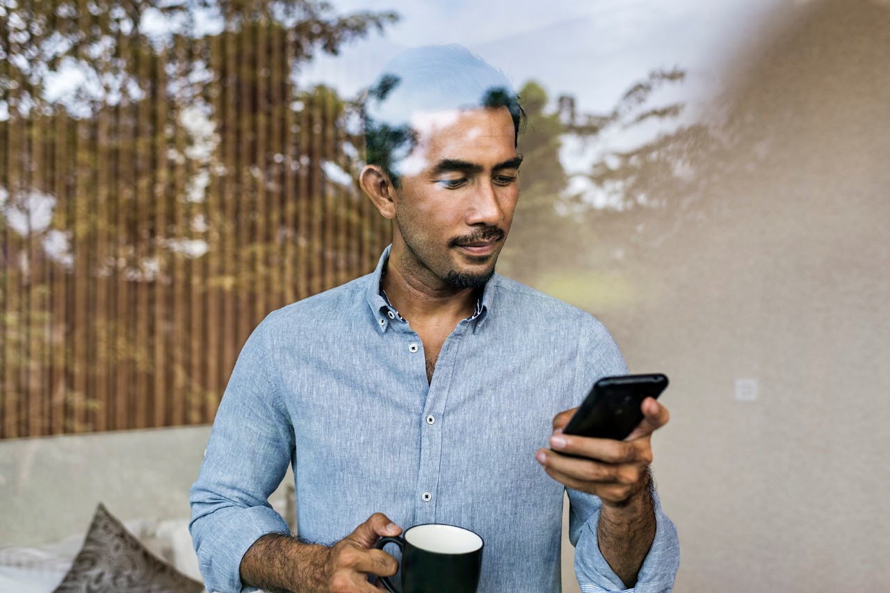 Asia, Indonesia, Jakarta, Asian businessman  working from home,  using smartphone and drinking coffee, while standing at bedroom  window of modern house